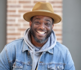 Geno Young is a black man wearing a blue jean jacket and a brown hat, smiling in front of a brick wall.
