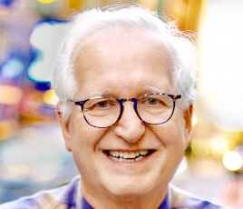 Jack has white hair and black glasses. He wears a blue and brown checked shirt against a blurred background.