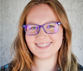 Justis Schneider is a girl with long brown hair, blue glasses, and wears a blue checkered shirt in front of a white background.