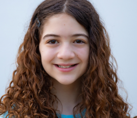 Millie Haber is a girl with long curly brown hair and wears a blue shirt with a black vest in front of a light background.