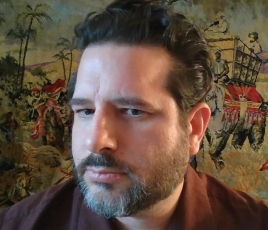 Paco José Madden has short dark hair and a dark beard and is wearing a red shirt in front of a multicolored background.