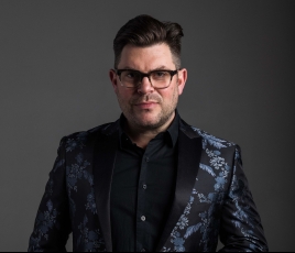 Sonny Paladino has short dark hair, wears black glasses, and wears a black jacket in front of a dark background.