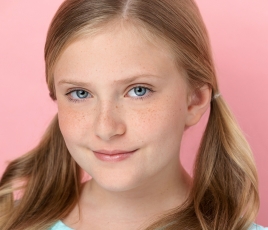 Kinley Stratton is a girl with long brown hair in pig tails and wears a blue shirt against a pink background.