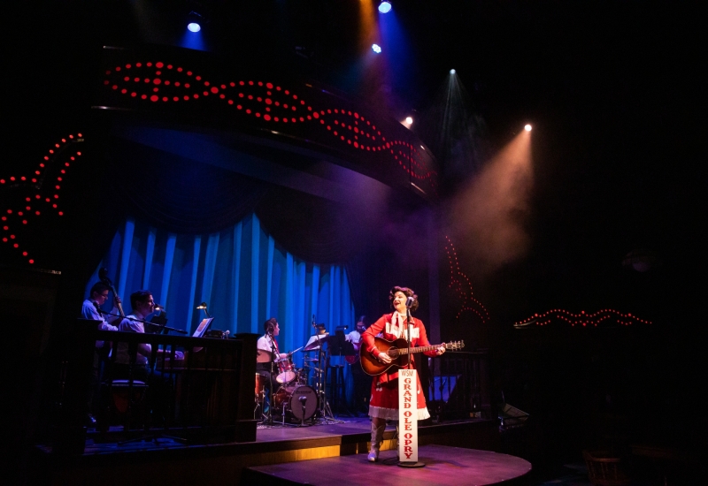Cassie Chilton as Patsy Cline in front of a band made up of Kevin White on piano, Ken Skaggs on the pedal steel guitar, Andrew Gonzalez on guitar, Nathaniel De La Cruz on bass, and Michelle Chin on drums.