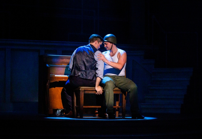 Alex Crossland sits at an upright piano leaning left, his back to the audience but facing EJ. EJ Dohring sits next to him on the bench, facing forward, with one hand on his shoulder representing a soldier in a war hat, white tank top, and green pants.